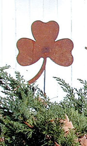Shamrock Rusted Garden Stake placed among evergreen shrubs, showcasing rustic charm for seasonal or year-round garden decoration.